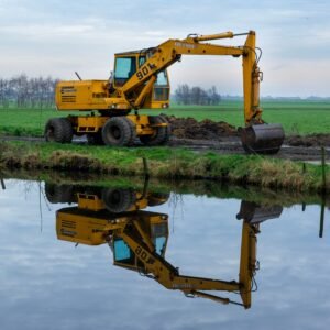 A yellow excavator working near a calm river with reflection in a serene rural landscape.